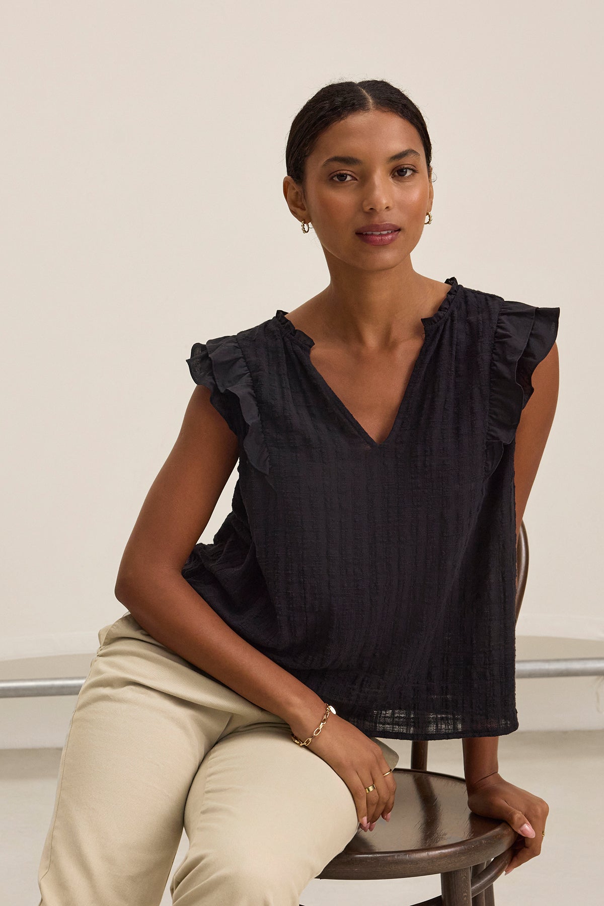 A woman sits on a chair in the EVA TEXTURED COTTON GAUZE TOP by Velvet by Graham & Spencer, featuring black ruffle sleeves and a v-neckline, paired with light pants, looking at the camera against a plain background.-39333220450497