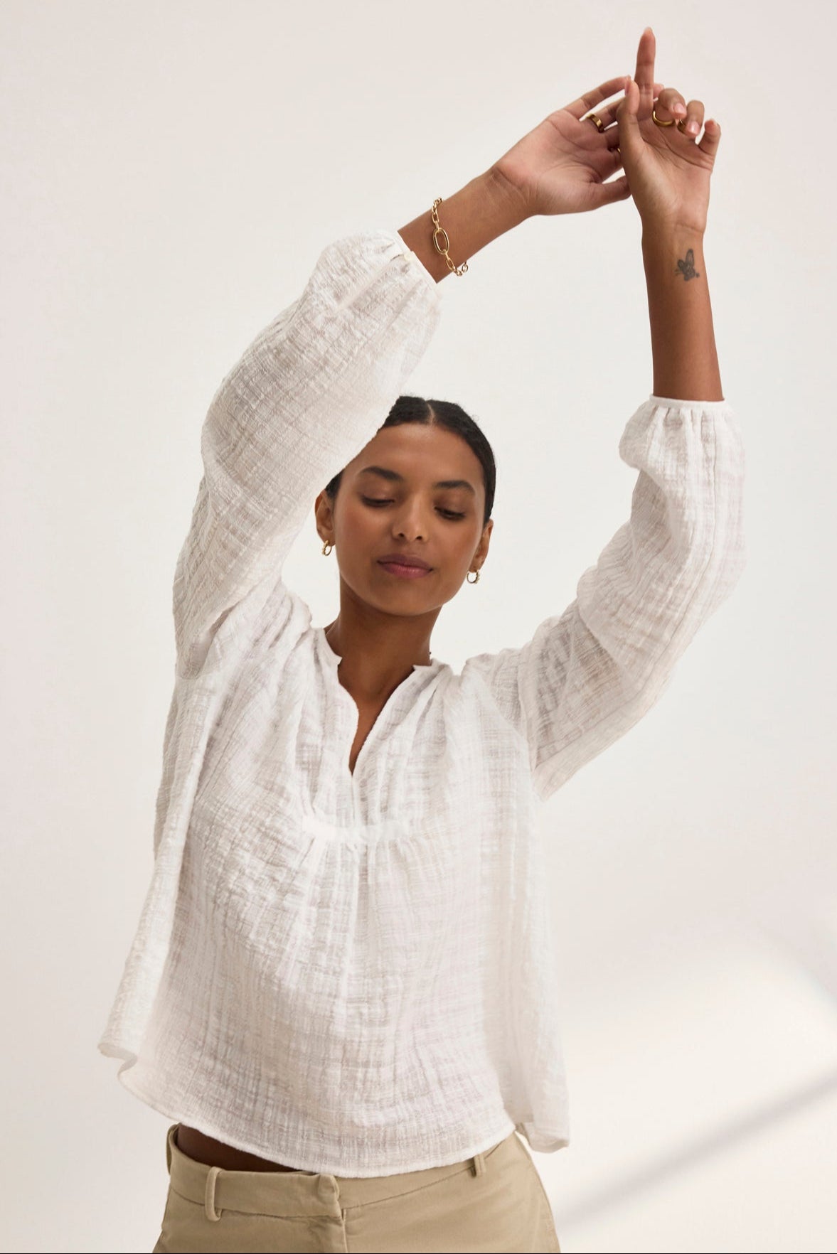 A person in a white KORA TEXTURED COTTON GAUZE TOP by Velvet by Graham & Spencer and beige pants, hands raised, eyes closed, showing a small tattoo on the right forearm.