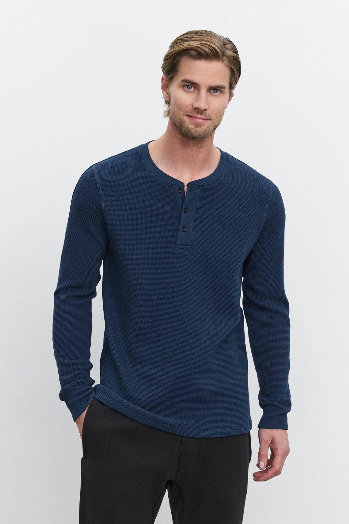 A man with light brown hair and a beard is wearing the DEX THERMAL HENLEY by Velvet by Graham & Spencer, a navy blue long-sleeve henley shirt with a 3-button placket, made of cotton thermal knit. He is also wearing black pants and standing against a plain white background.-37893766709441