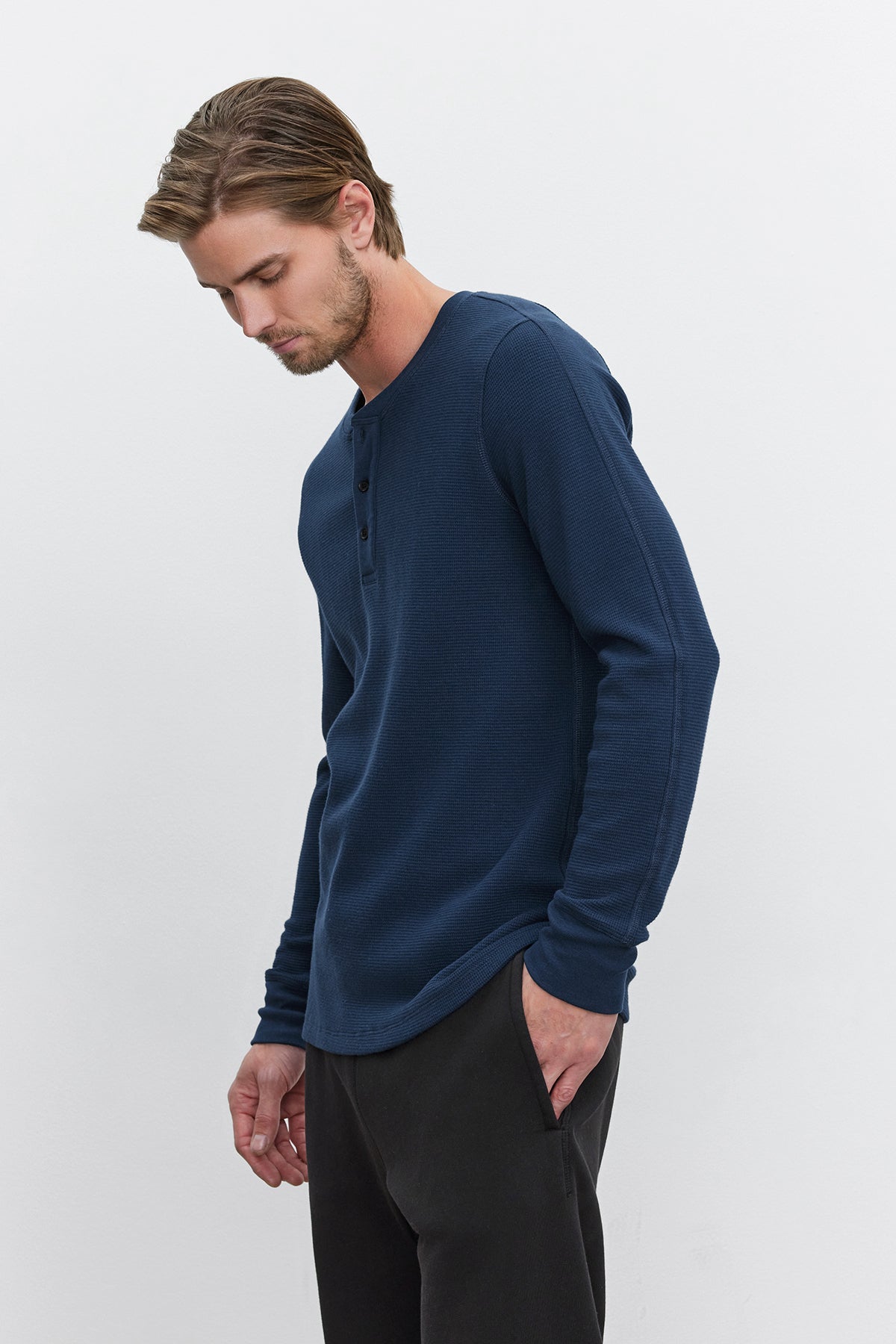 A man with light brown hair, dressed in a dark blue Velvet by Graham & Spencer DEX THERMAL HENLEY featuring a 3-button placket and dark pants, looks down to his right with his left hand in his pocket against a plain white background.