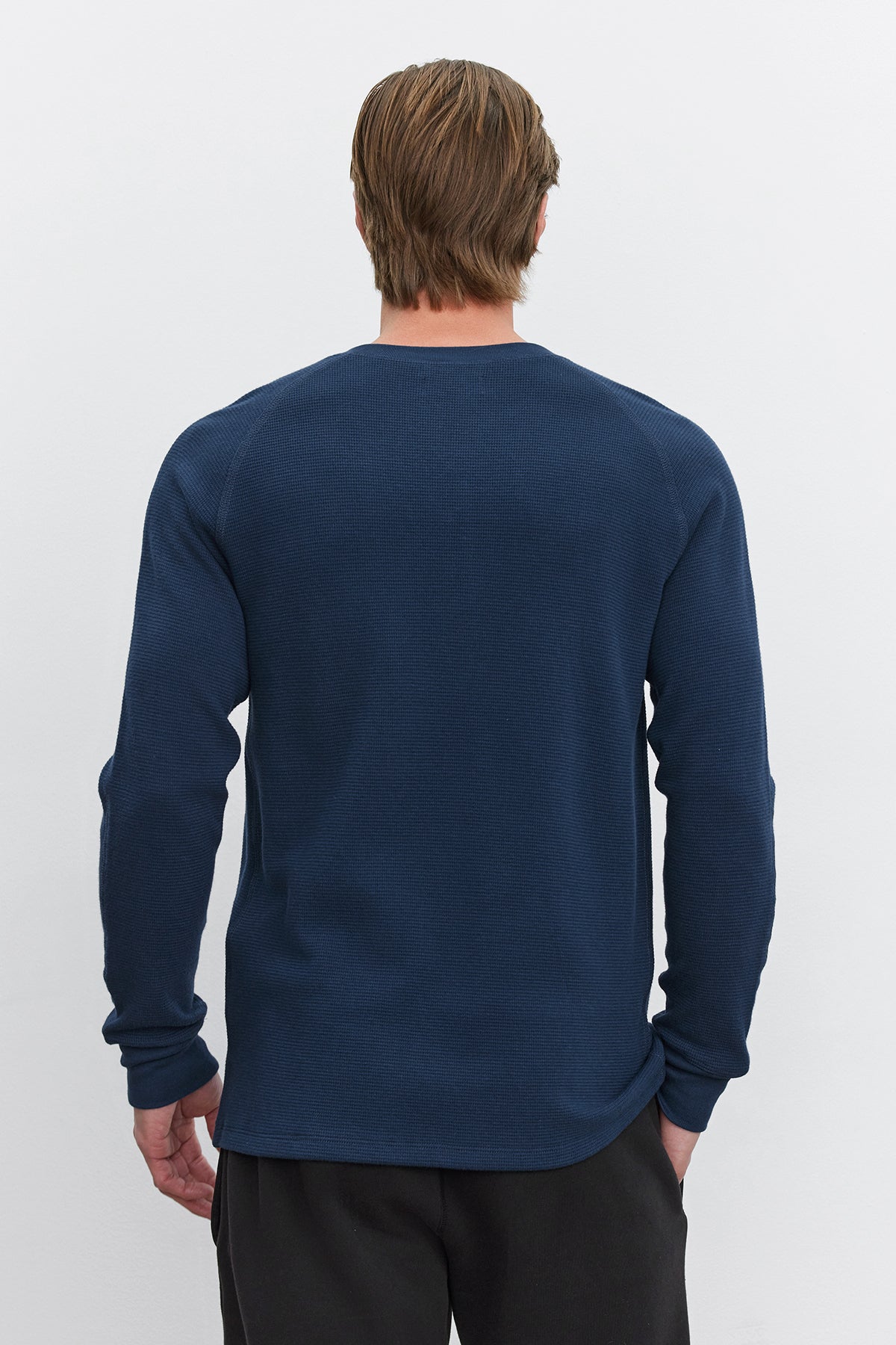 A person with medium-length hair wearing a long-sleeved, blue DEX THERMAL HENLEY with a 3-button placket from Velvet by Graham & Spencer and dark pants is shown from the back against a plain white background.