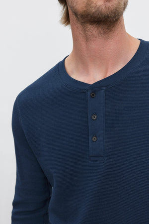 A man wears the DEX THERMAL HENLEY by Velvet by Graham & Spencer, a blue, long-sleeve henley shirt featuring a 3-button placket. He is shown from the shoulders to just above the nose. The cotton thermal knit makes it perfect for layering.