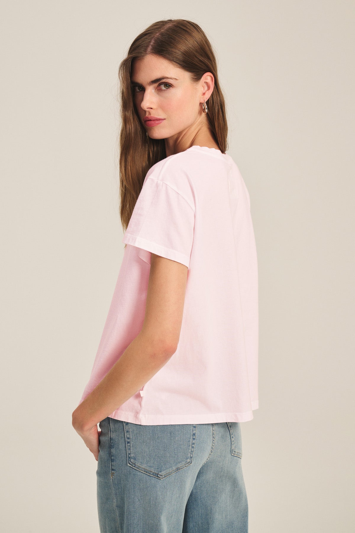 A woman with long brown hair wears the TIA TEE by Velvet by Graham & Spencer—a light pink cotton jersey crewneck tee—paired with light blue jeans, standing sideways and looking over her shoulder; a timeless wardrobe essential.-41044148420801