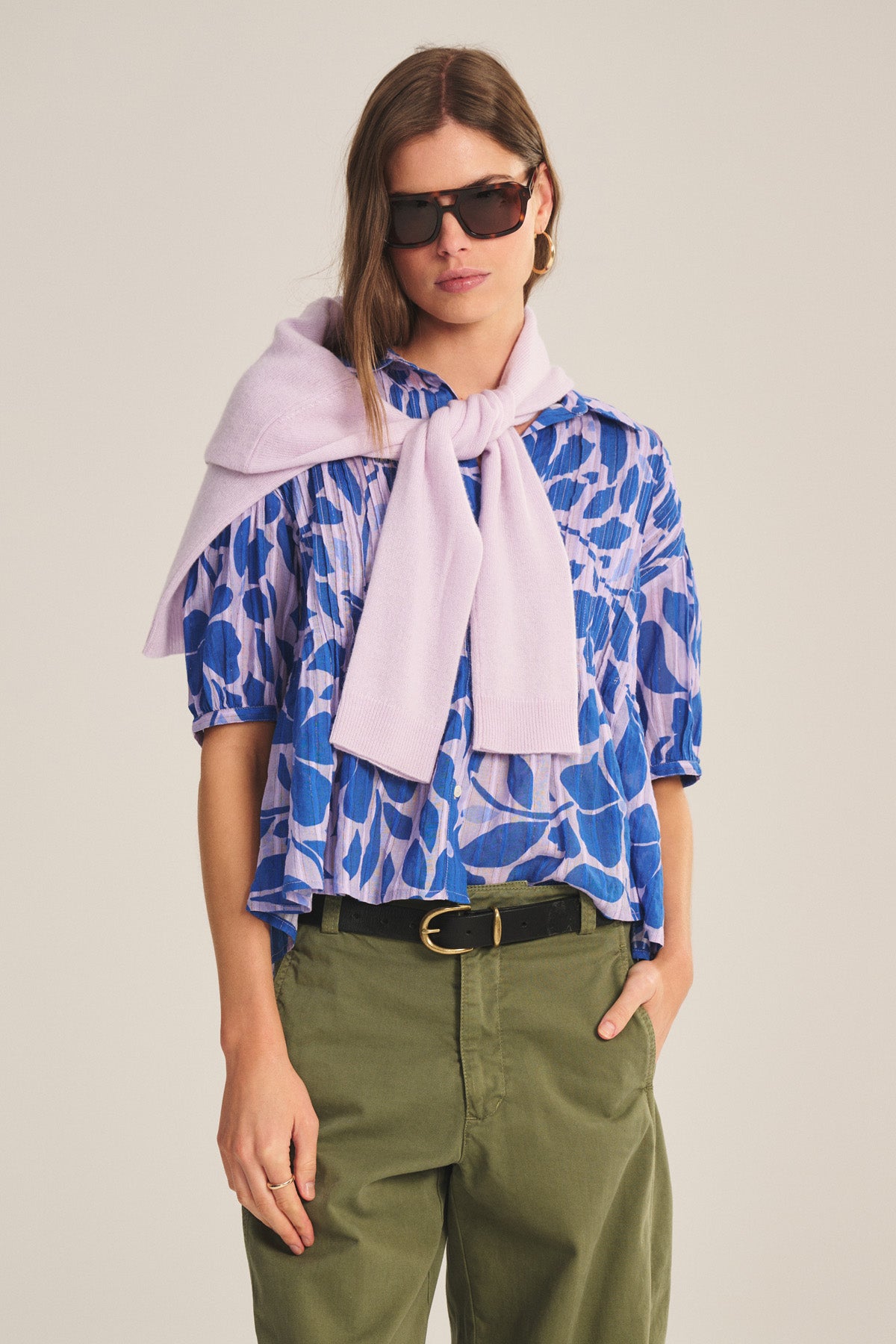 A woman wears the TILDA TOP by Velvet by Graham & Spencer, sunglasses, olive green pants, and a light purple sweater draped over her shoulders, standing with one hand in her pocket.-40808749039809