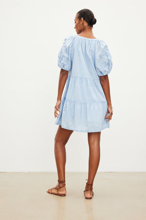 A person wearing the CHRISSY DRESS by Velvet by Graham & Spencer—a light blue, short-sleeved, cotton dress with a v-neckline and tiered skirt, adorned with floral embroidery on the sleeves—facing away from the camera. They are accessorizing with brown sandals and gold hoop earrings.