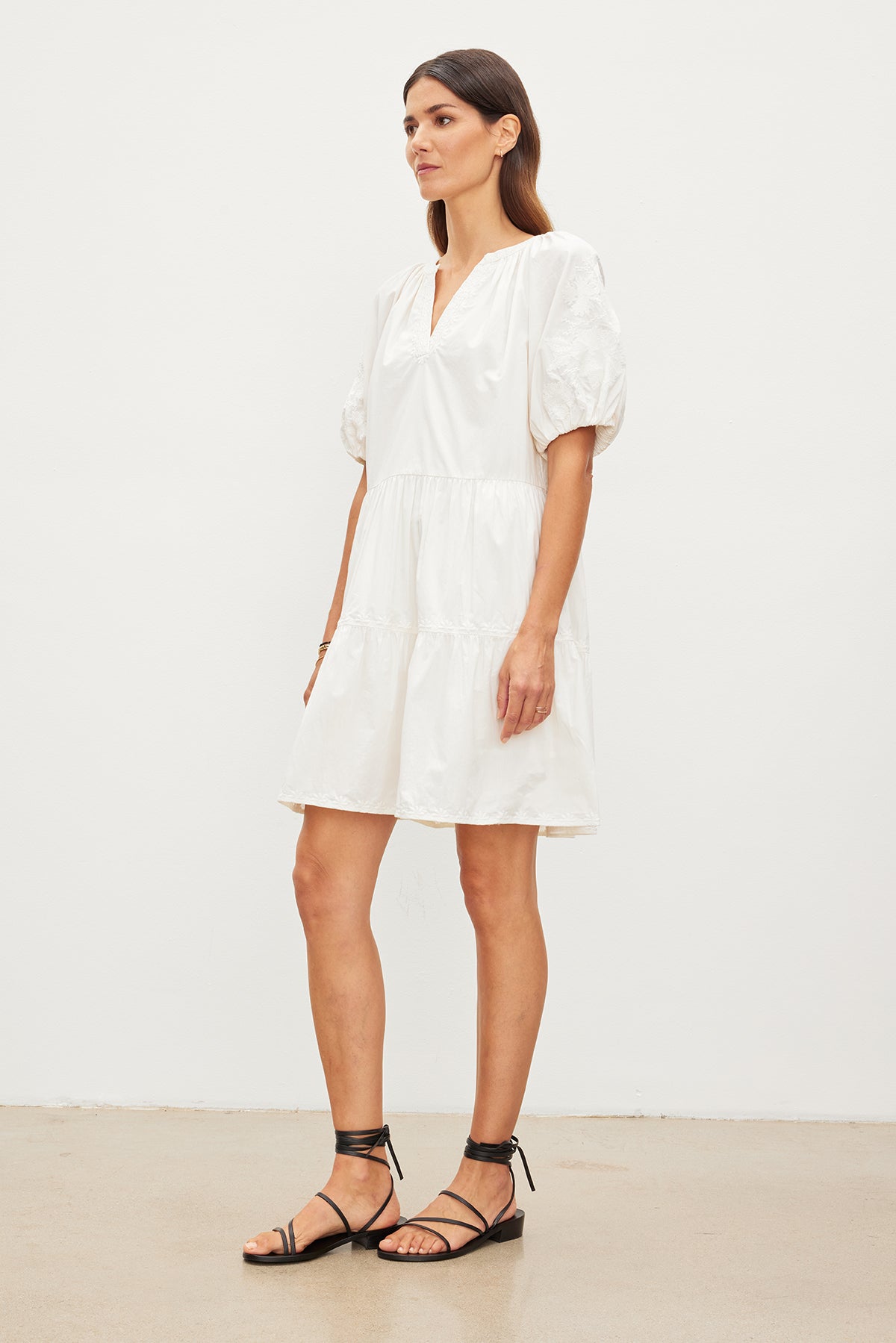 A woman wearing the CHRISSY DRESS by Velvet by Graham & Spencer, characterized by its white color, short sleeves, and v-neckline, stands against a plain white background while sporting black strappy sandals.-35955557499073