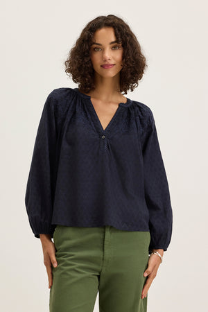 A woman with curly brown hair models the Velvet by Graham & Spencer CARINA EMBROIDERED TOP— a navy, long-sleeve, V-neck blouse—paired with green pants, standing against a plain light background.