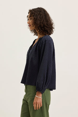 A woman with curly hair stands sideways against a plain light background, wearing the Velvet by Graham & Spencer CARINA EMBROIDERED TOP in navy with tonal embroidery and olive green pants.