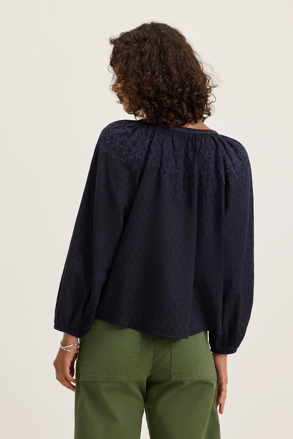 A person with curly hair wears the CARINA EMBROIDERED TOP by Velvet by Graham & Spencer, a lightweight cotton piece with tonal embroidery, paired with green pants and shown from the back against a plain background.-39333218877633