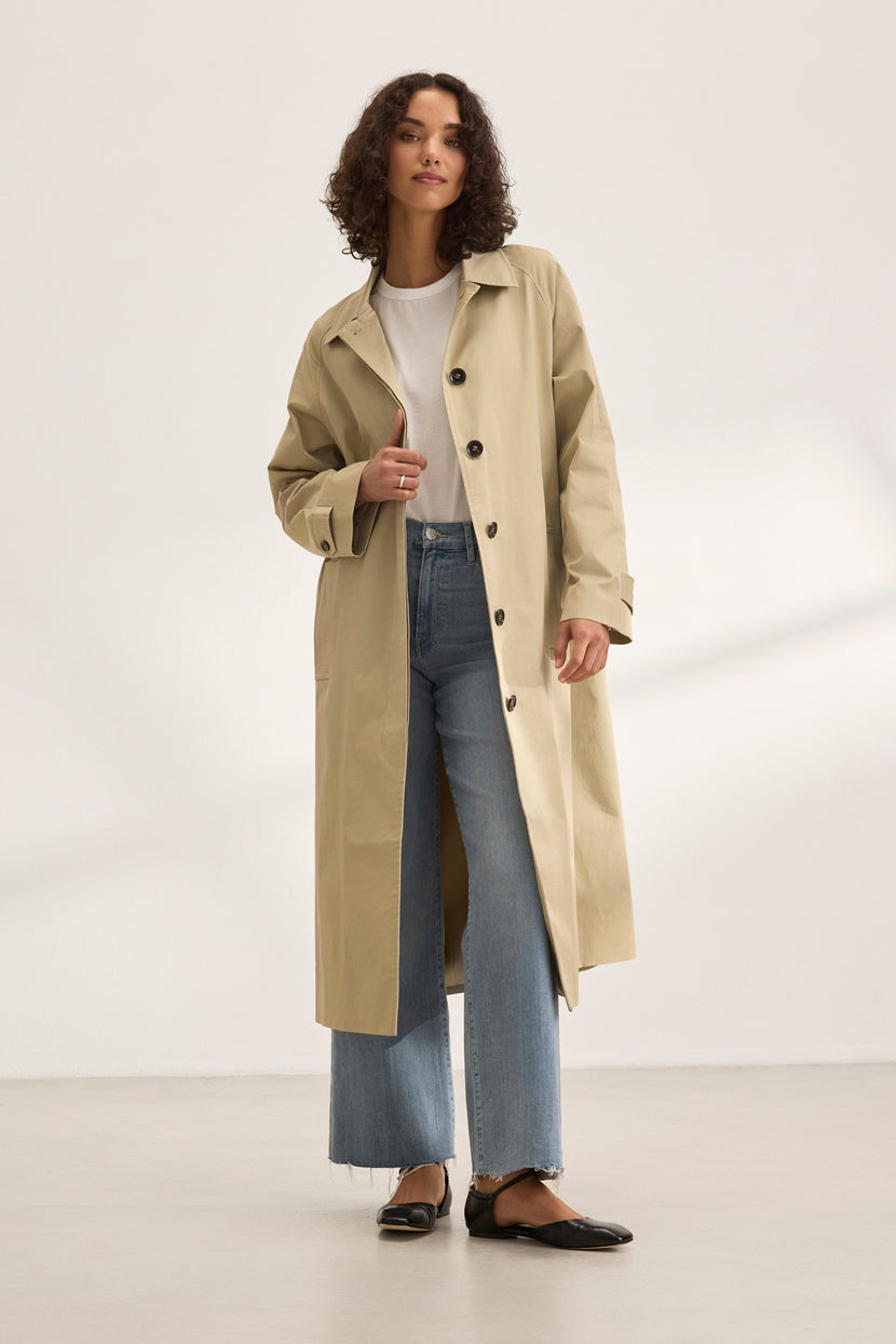 Against a plain backdrop, a person models the RAHEAL TRENCH COAT by Velvet by Graham & Spencer. The timeless beige coat with its cotton-elastane shell is paired with a white t-shirt, blue jeans, and black shoes, exemplifying versatile styling for any occasion.
