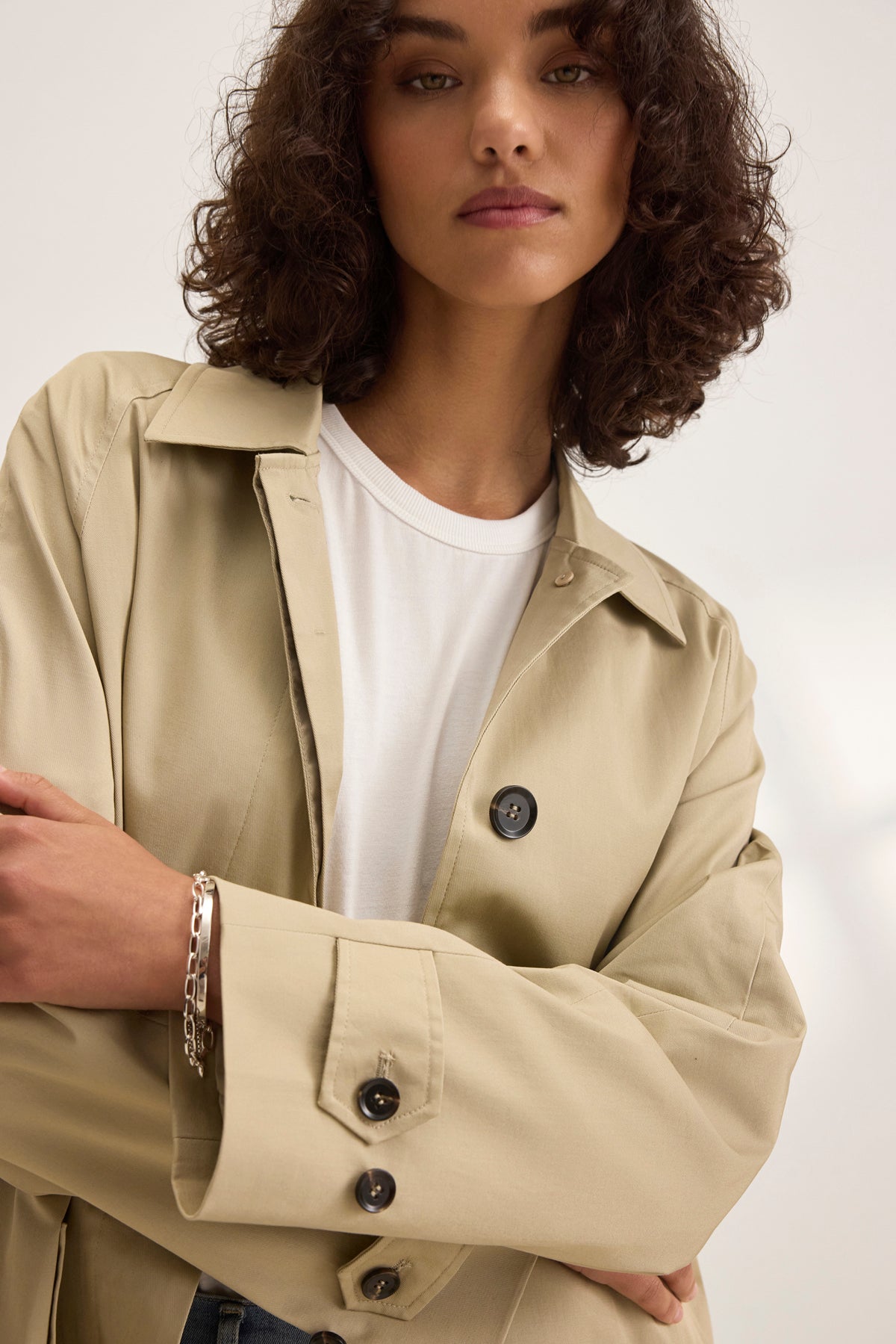 The curly-haired individual stands confidently, arms crossed, wearing the RAHEAL TRENCH COAT by Velvet by Graham & Spencer. The beige coat features a detachable belt over their crisp white shirt.-38819770728641