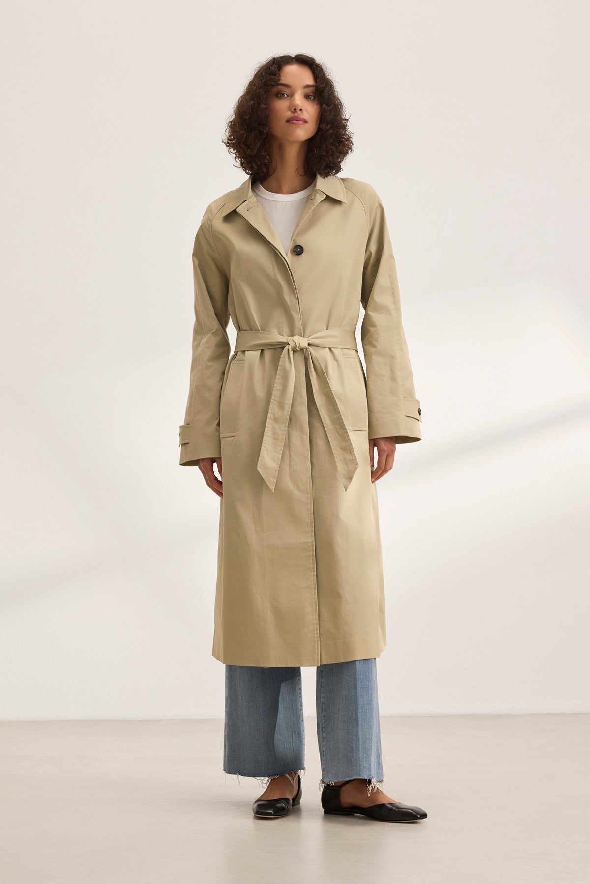 A person models the versatile RAHEAL TRENCH COAT by Velvet by Graham & Spencer. They pair it with light blue jeans and black shoes against a plain backdrop, highlighting the beige cotton-elastane coat's stylish design with a belt.