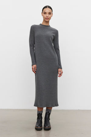 A woman is standing against a plain white background, wearing the LIZ RIBBED DRESS, a long-sleeved, ankle-length gray knit dress by Velvet by Graham & Spencer, paired with black boots.