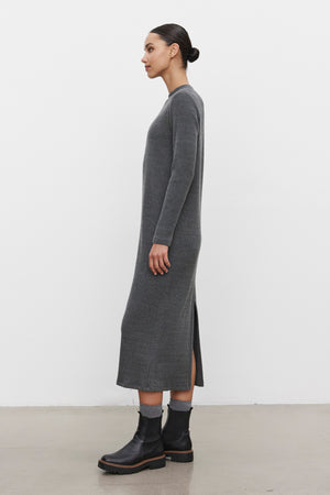 A person stands in profile wearing the LIZ RIBBED DRESS from Velvet by Graham & Spencer, a long, gray cotton blend dress featuring a crew neckline and long sleeves. They pair it with black boots and have their hair tied back, standing against a plain white background.