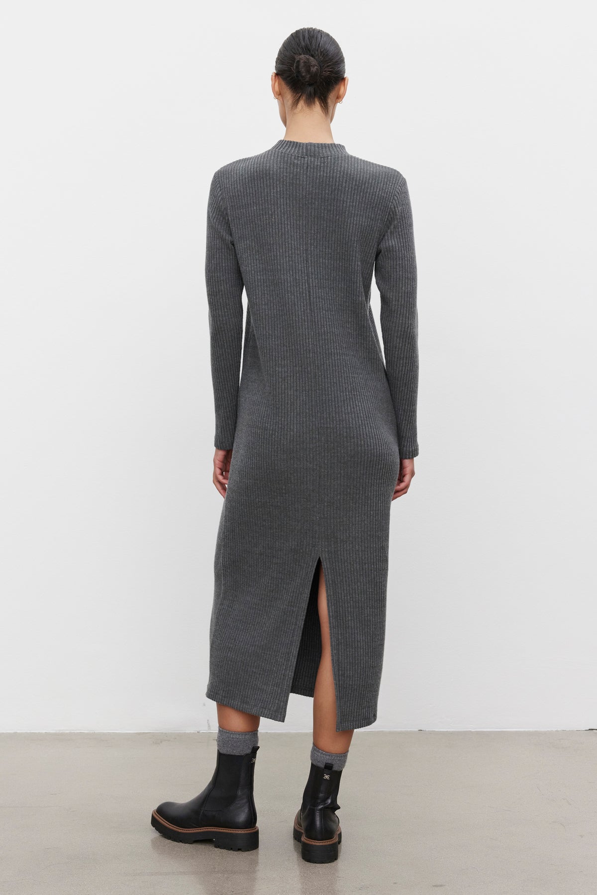 A person stands against a white background, wearing the LIZ RIBBED DRESS from Velvet by Graham & Spencer. The long-sleeved, ankle-length grey cotton blend dress features a back slit and is paired with black ankle boots. The ribbed fabric adds texture while the crew neckline completes the elegant look as the person faces away from the camera.-37856329367745