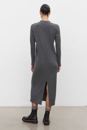 A person stands against a white background, wearing the LIZ RIBBED DRESS from Velvet by Graham & Spencer. The long-sleeved, ankle-length grey cotton blend dress features a back slit and is paired with black ankle boots. The ribbed fabric adds texture while the crew neckline completes the elegant look as the person faces away from the camera.