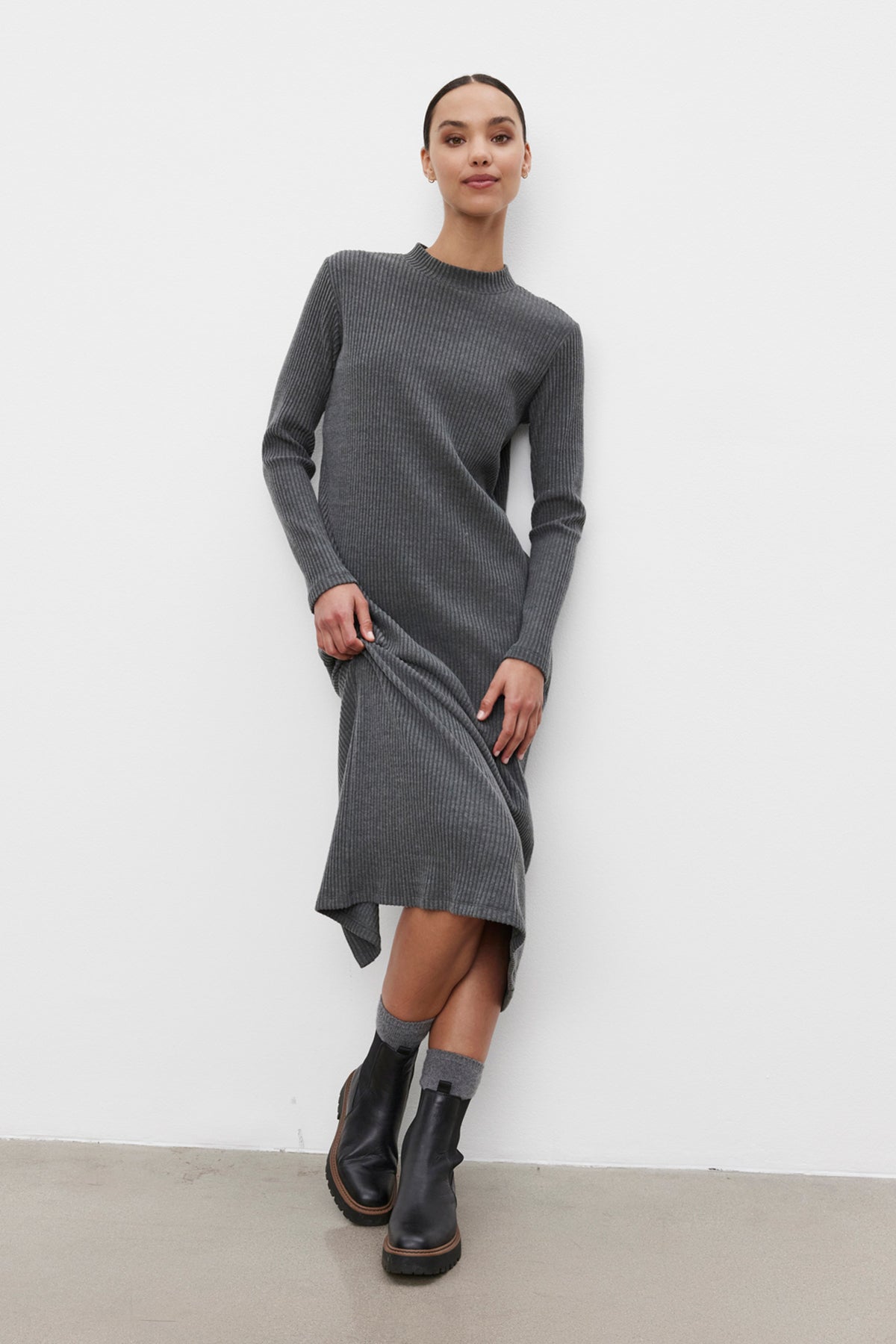 A person poses in a knee-length, long-sleeved LIZ RIBBED DRESS by Velvet by Graham & Spencer with a crew neckline and black ankle boots, standing against a white wall.-37856329334977