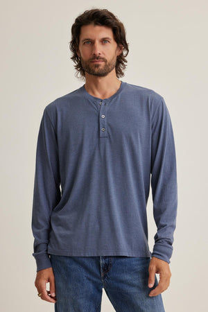A bearded man with medium-length hair stands against a plain background, wearing the long-sleeved DESI HENLEY in blue by Velvet by Graham & Spencer, paired with jeans.