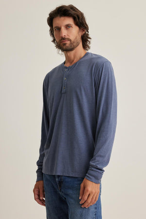 A man with long hair and a beard in a blue Velvet by Graham & Spencer DESI HENLEY and jeans stands against a plain background.