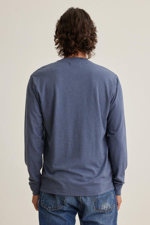 A medium-haired individual models the DESI HENLEY by Velvet by Graham & Spencer, wearing a blue long-sleeve shirt and jeans, facing away from the camera against a plain background.