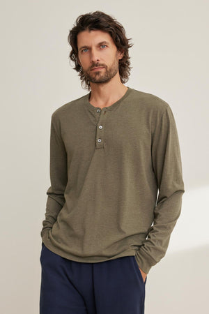 Man with medium-length hair and beard stands against a beige background wearing Velvet by Graham & Spencer's olive green DESI HENLEY long-sleeve shirt and dark pants.