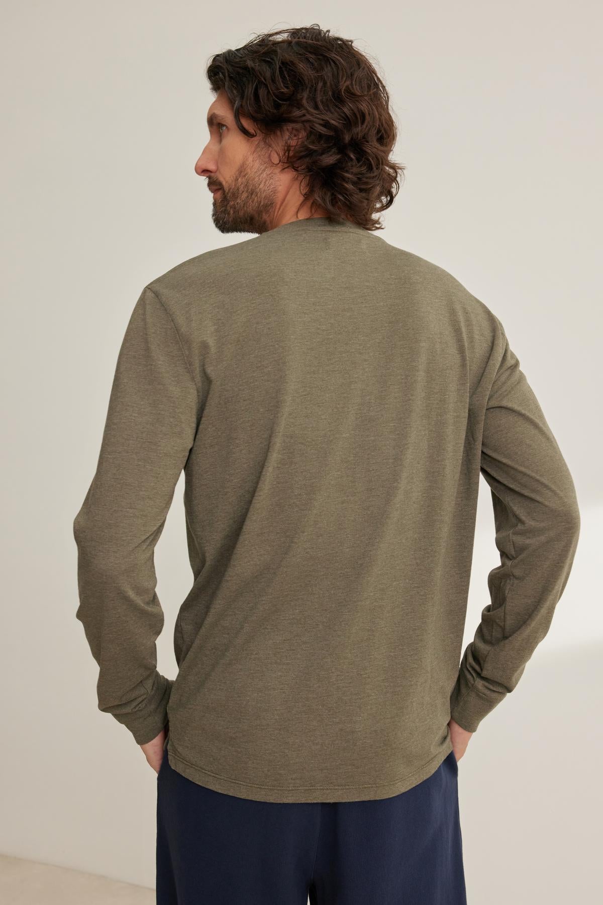 A man with wavy hair is standing, facing slightly to the side, wearing a Velvet by Graham & Spencer DESI HENLEY long-sleeved olive green shirt and dark pants.