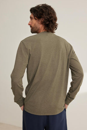A man with wavy hair is standing, facing slightly to the side, wearing a Velvet by Graham & Spencer DESI HENLEY long-sleeved olive green shirt and dark pants.