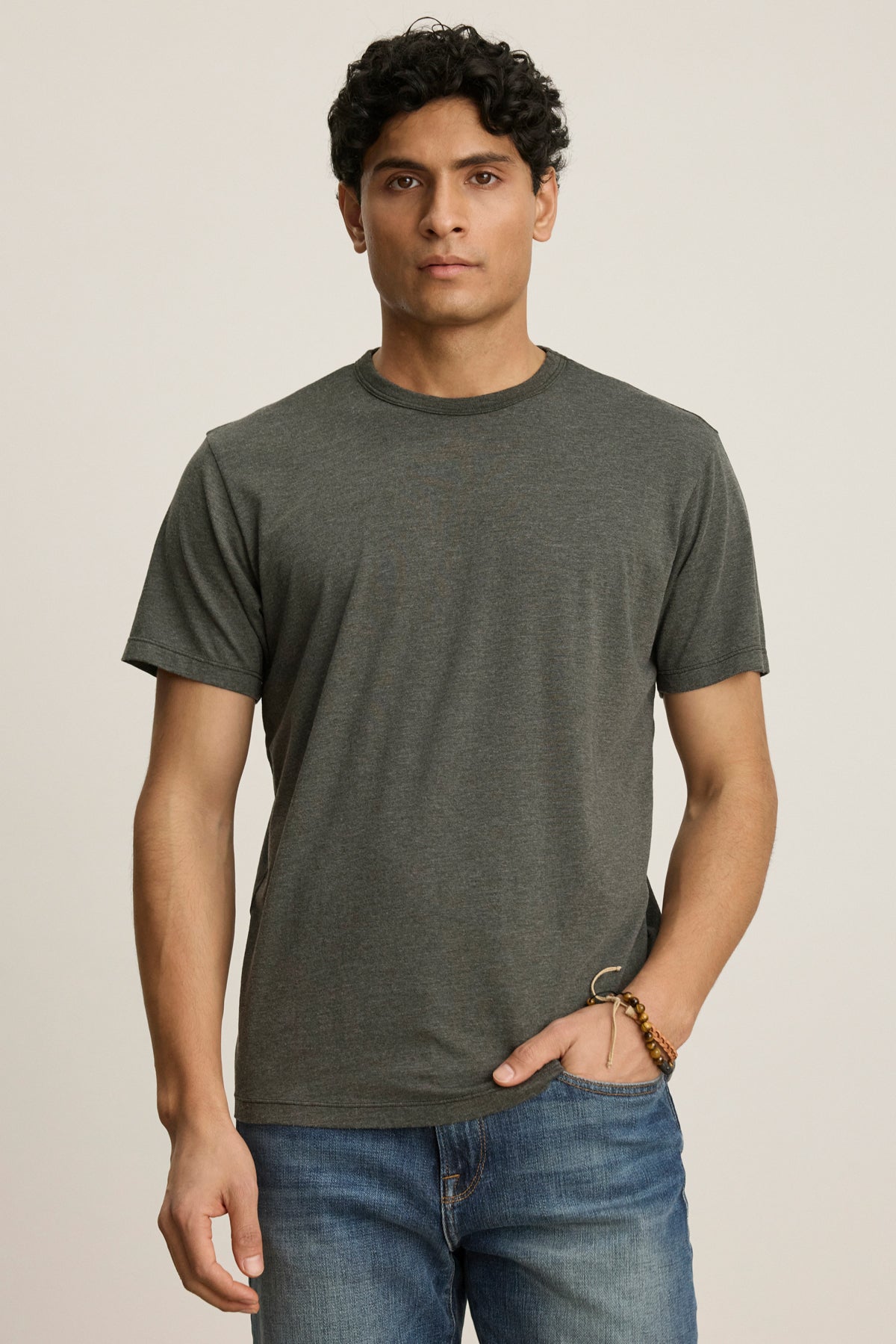 A man with short curly hair and a neutral expression wears the ZANDER TEE by Velvet by Graham & Spencer—a plain dark gray t-shirt with a ribbed crew neckline—paired with blue jeans, standing against a light background.-39446054928577