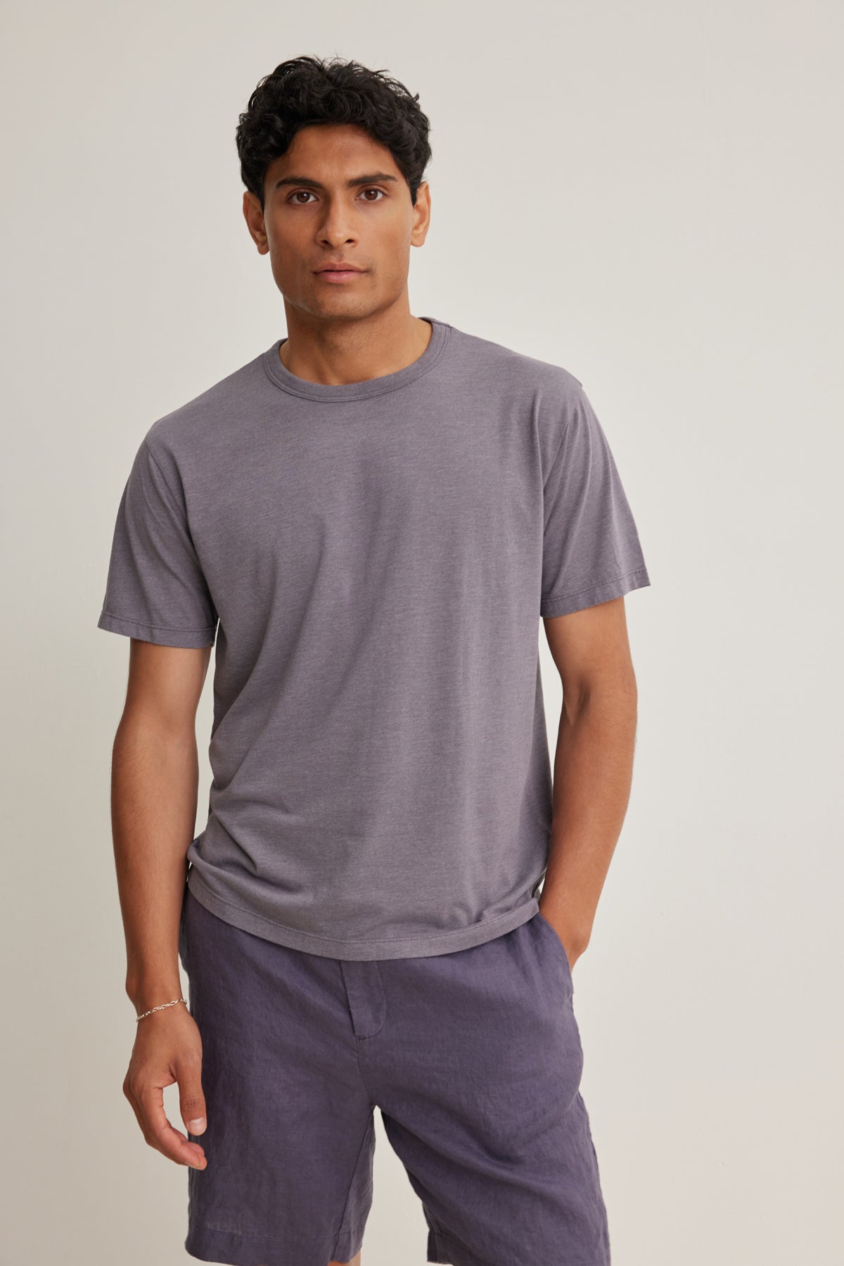 A man stands against a light background wearing the Velvet by Graham & Spencer ZANDER TEE, a relaxed fit short-sleeved gray t-shirt, paired with dark gray shorts for an effortlessly wearable everyday look.