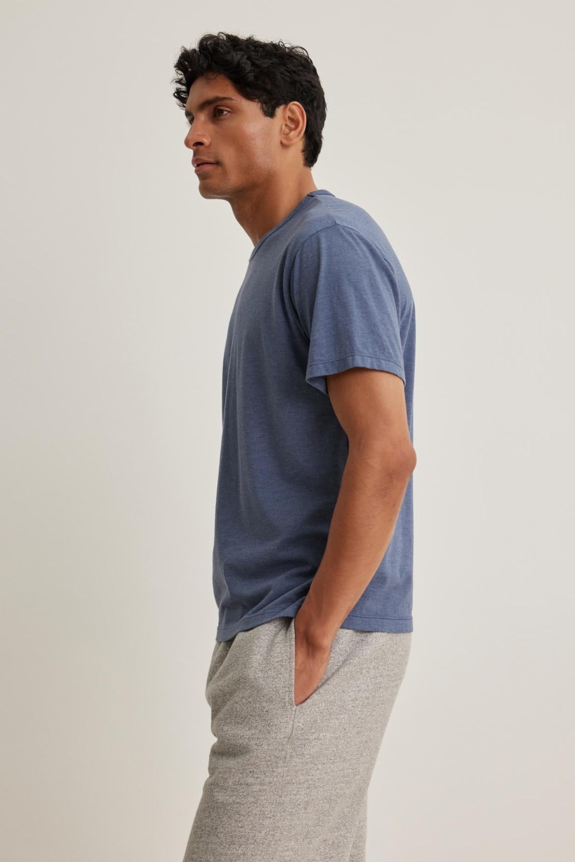 A person wearing a Velvet by Graham & Spencer ZANDER TEE in blue and gray pants stands with hands in pockets, looking left against a plain light background.