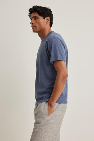 A person wearing a Velvet by Graham & Spencer ZANDER TEE in blue and gray pants stands with hands in pockets, looking left against a plain light background.