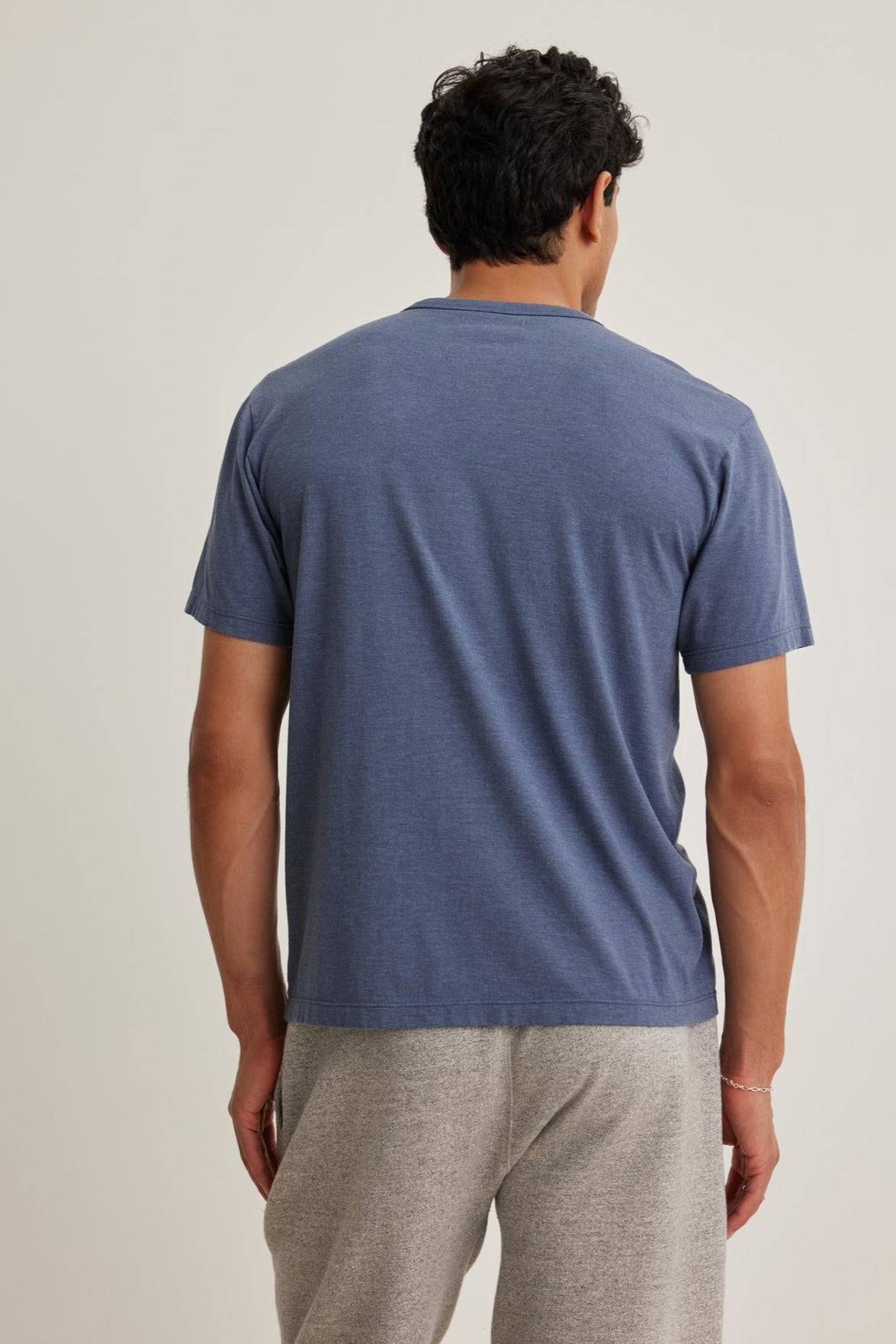A man with curly hair stands back to the camera, wearing a blue ZANDER TEE by Velvet by Graham & Spencer and gray pants.