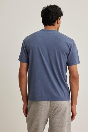 A man with curly hair stands back to the camera, wearing a blue ZANDER TEE by Velvet by Graham & Spencer and gray pants.