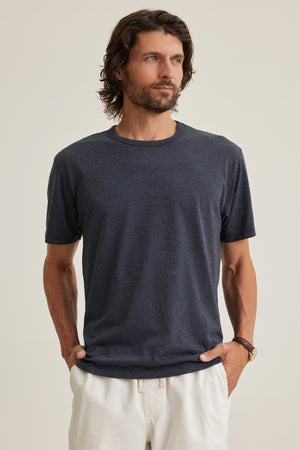 A man with wavy brown hair and a beard wears the Velvet by Graham & Spencer ZANDER TEE, a relaxed fit dark gray cotton-modal blend t-shirt, and white pants, standing with his hands in his pockets against a plain background.