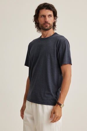 Man wearing the ZANDER TEE by Velvet by Graham & Spencer—a dark gray, relaxed fit short-sleeve t-shirt—paired with white pants and set against a plain light background, ideal for effortless everyday style.