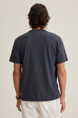 A person with medium-length brown hair faces away, wearing the Velvet by Graham & Spencer ZANDER TEE in dark relaxed fit and white pants against a plain background—ideal for everyday wearability.