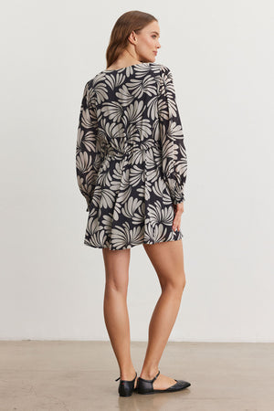 Wearing the TATTI DRESS by Velvet by Graham & Spencer, a person in a black and white floral design with a V-neckline is shown from behind, emphasizing the back details of the printed viscose fabric. The look is complemented by black flat shoes on a plain floor.