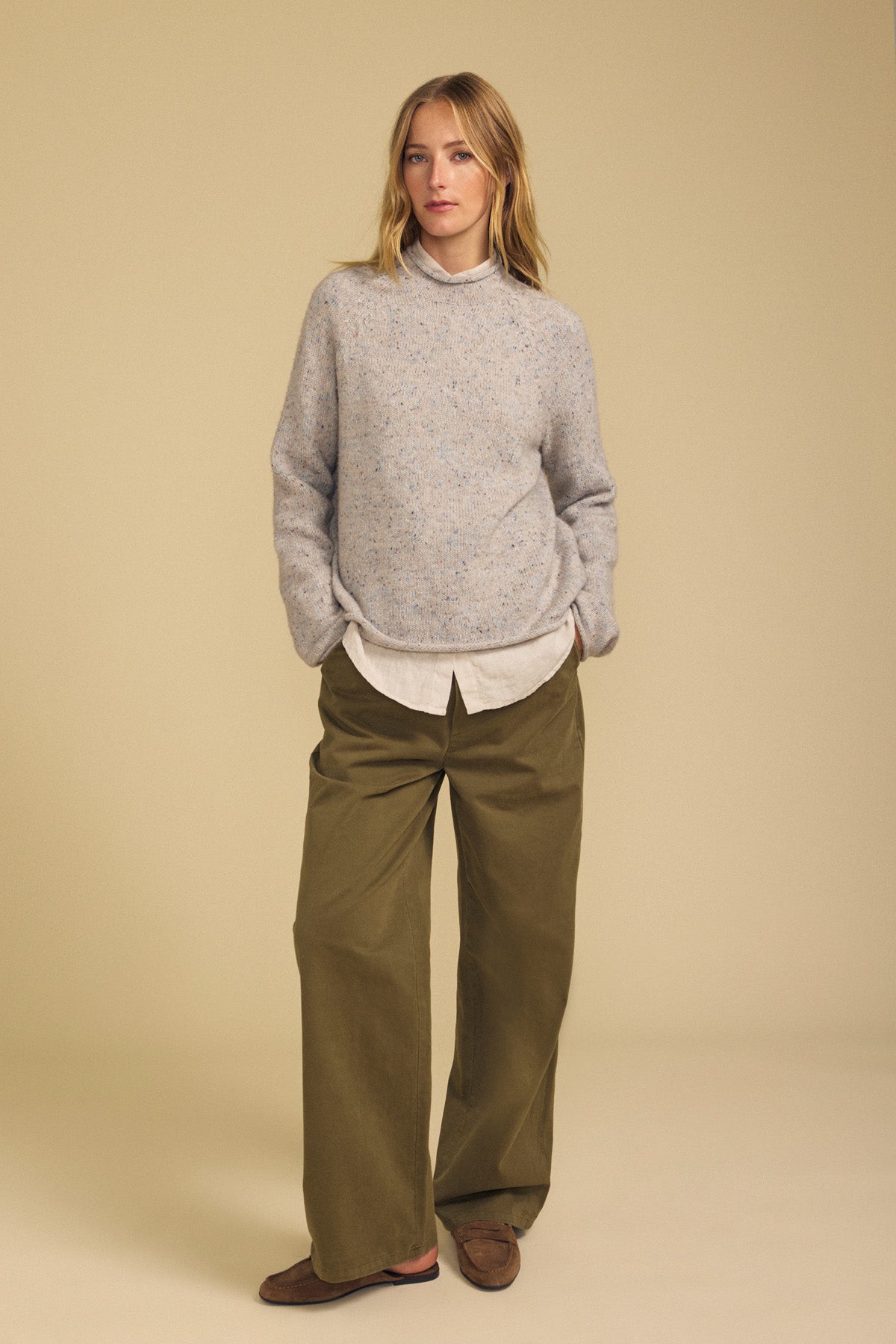 A person stands against a beige background, wearing the Velvet by Graham & Spencer JOELLE SWEATER—a light gray speckled mock neck style—layered over a white shirt, paired with olive green wide-leg pants and brown shoes. Their hands are in their pockets.-42063767699649
