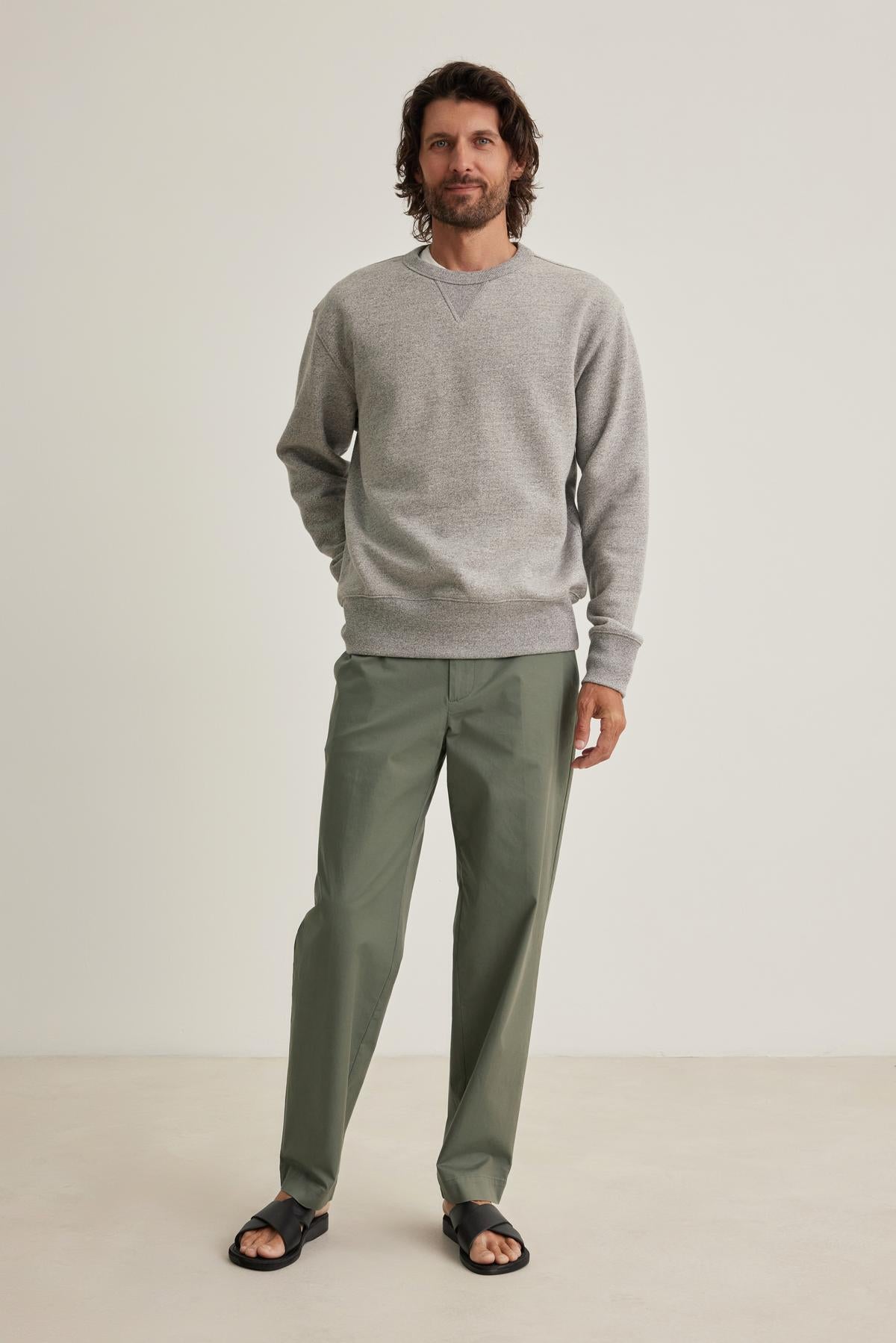 A man with long hair stands against a plain background, wearing a gray HAMES SWEATSHIRT by Velvet by Graham & Spencer, green pants, and black sandals.-38643296600257