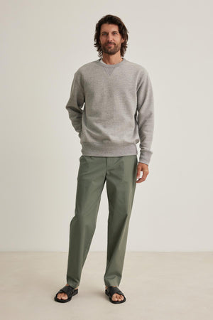 A man with long hair stands against a plain background, wearing a gray HAMES SWEATSHIRT by Velvet by Graham & Spencer, green pants, and black sandals.