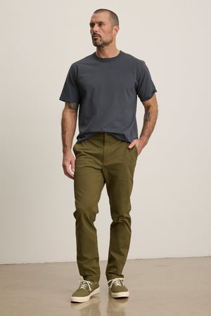 A man stands indoors against a plain light-colored wall, wearing a dark gray t-shirt, Velvet by Graham & Spencer MADDOX PANT in olive green stretch cotton twill, and beige sneakers.