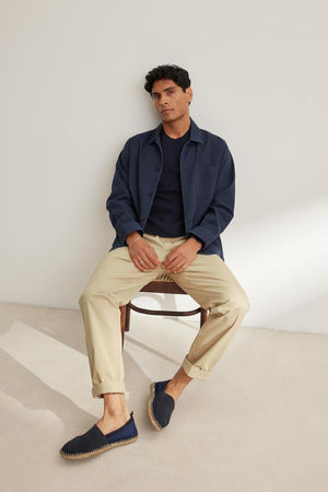 A person sits on a chair against a plain wall, wearing a navy jacket, dark shirt, Velvet by Graham & Spencer's STONE PANT in beige, and blue slip-on shoes.