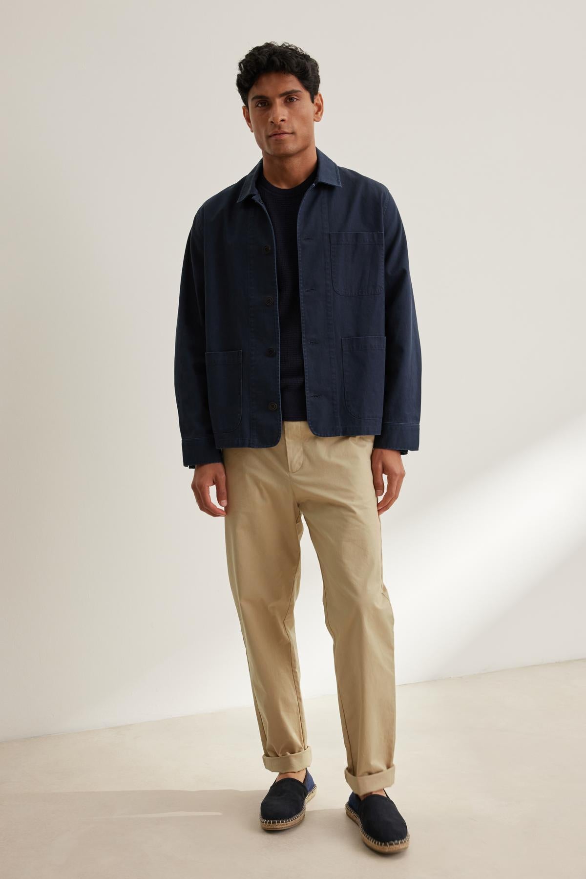 A man stands against a plain background wearing a navy jacket, black shirt, STONE PANT by Velvet by Graham & Spencer in beige, and dark slip-on shoes.-38643359842497