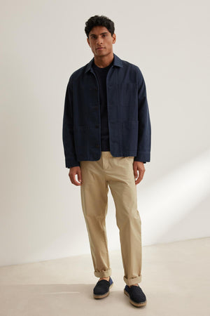 A man stands against a plain background wearing a navy jacket, black shirt, STONE PANT by Velvet by Graham & Spencer in beige, and dark slip-on shoes.