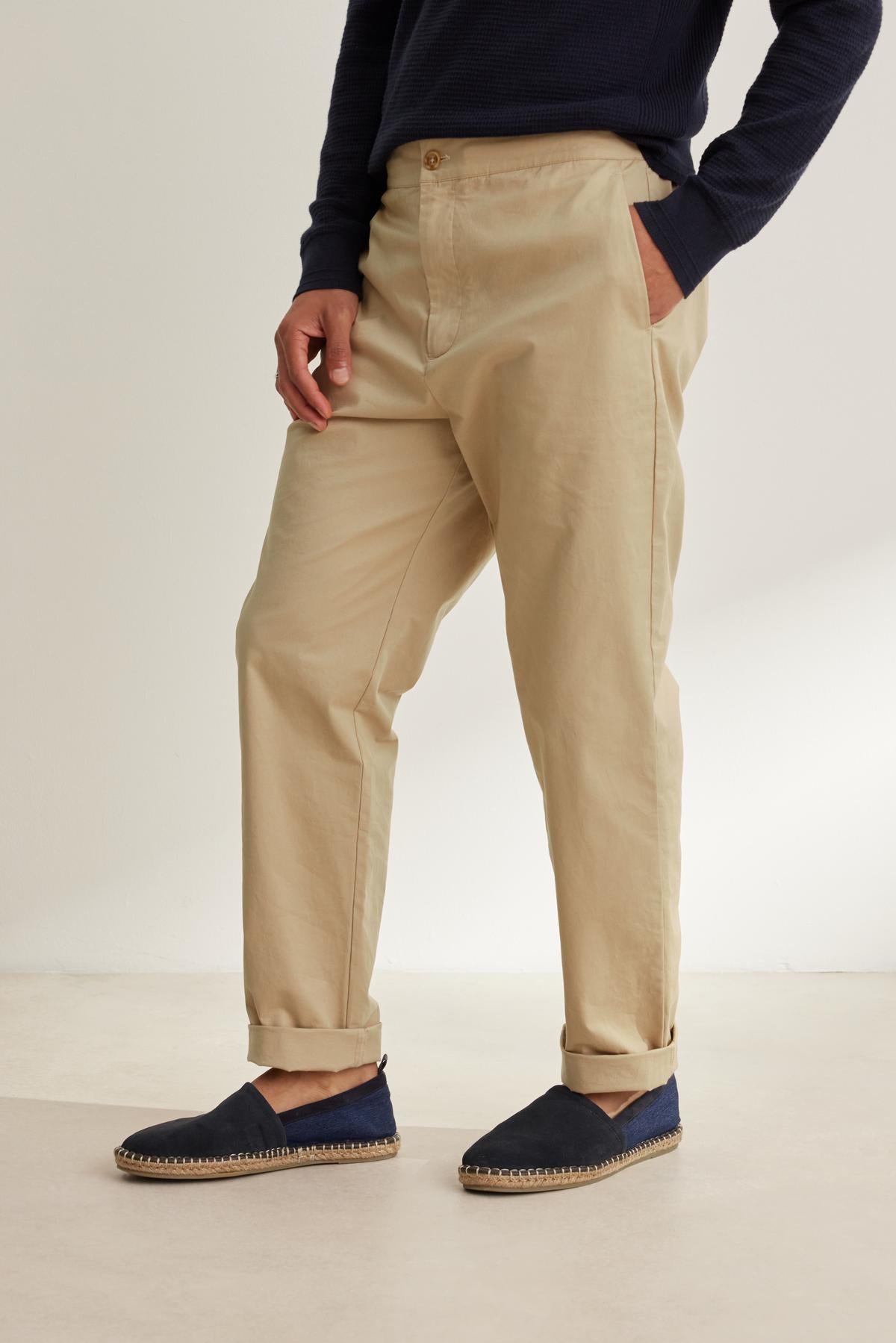 A person wearing a dark sweater, STONE PANT by Velvet by Graham & Spencer, and blue slip-on shoes with a tan sole stands with one hand in their pocket.-38643359482049