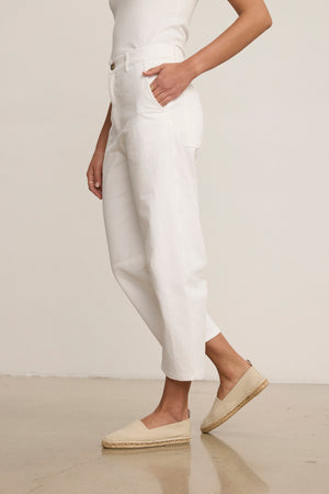 A person stands on a polished floor wearing a white sleeveless top, Velvet by Graham & Spencer’s ALEXIS PANT in white cotton twill with knee darting, and beige espadrille flats.