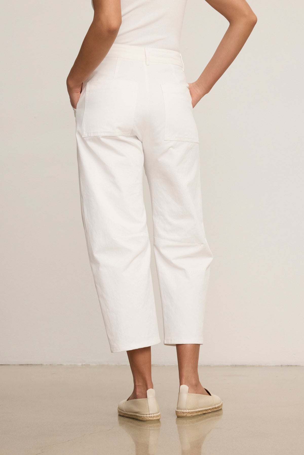 A person wearing the ALEXIS PANT by Velvet by Graham & Spencer, featuring white cotton twill and knee darting, stands with hands in pockets and faces away from the camera, styled with a sleeveless top and beige espadrilles.-39119701409985