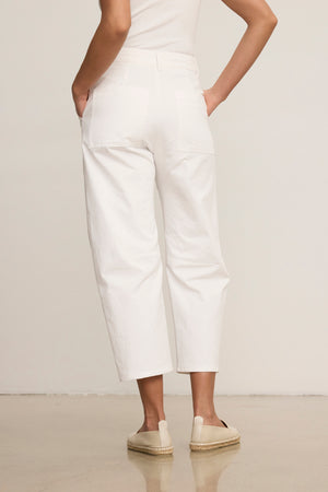 A person wearing the ALEXIS PANT by Velvet by Graham & Spencer, featuring white cotton twill and knee darting, stands with hands in pockets and faces away from the camera, styled with a sleeveless top and beige espadrilles.