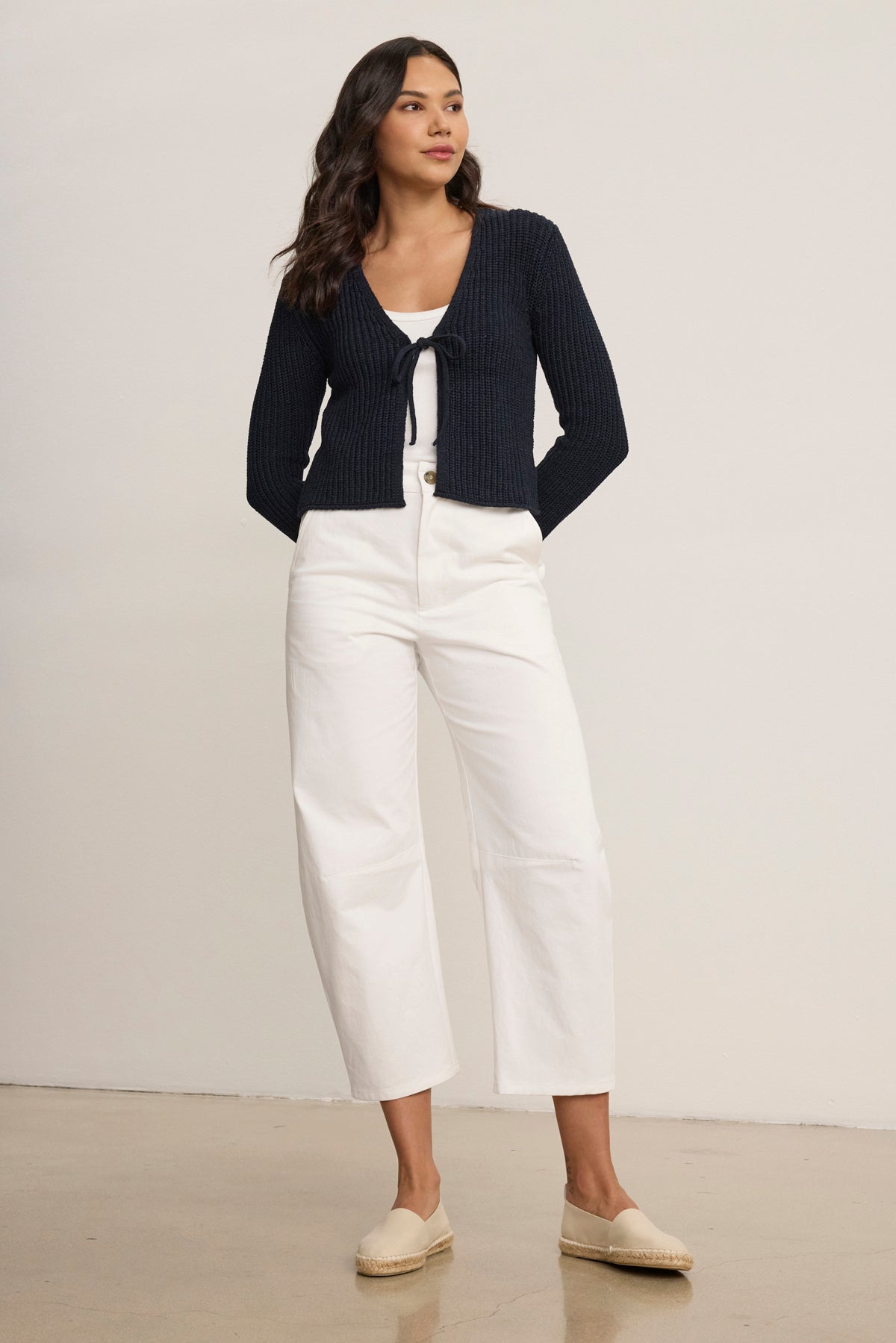 A woman stands indoors wearing a navy blue tie-front cardigan, a white top, Velvet by Graham & Spencer's ALEXIS PANT, and beige espadrilles.-39119701344449