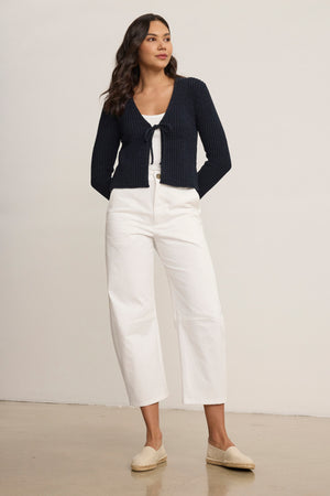 A woman stands indoors wearing a navy blue tie-front cardigan, a white top, Velvet by Graham & Spencer's ALEXIS PANT, and beige espadrilles.